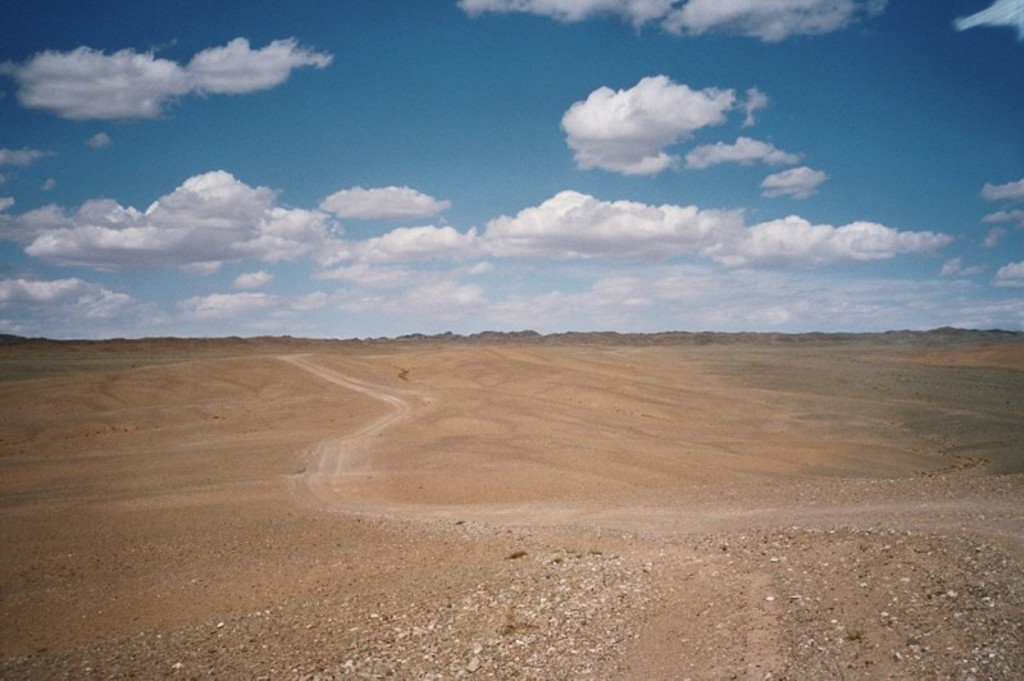Uitgestrekte steppe in Mongolië met zandpad onder blauwe lucht met wolken.