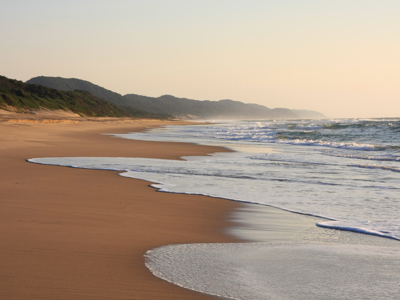 Zandstrand aan de Zuid-Afrikaanse kust met golven en duinen tijdens zonsondergang op de route Kaapstad–Johannesburg.