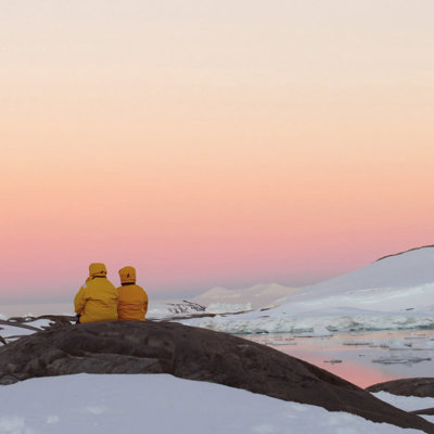 Twee reizigers in gele jassen kijken uit over ijsbergen en besneeuwde kust in Antarctica bij zonsondergang.
