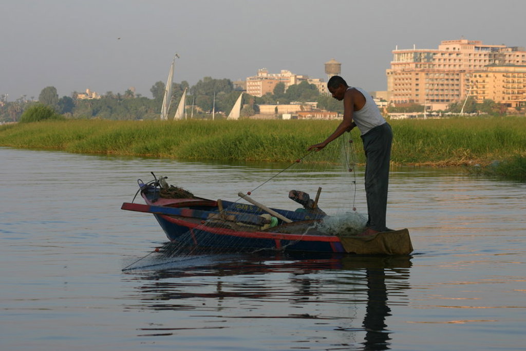 Visser in boot op de Nijl bij Luxor, Egypte, met stad en zeilboten op de achtergrond.