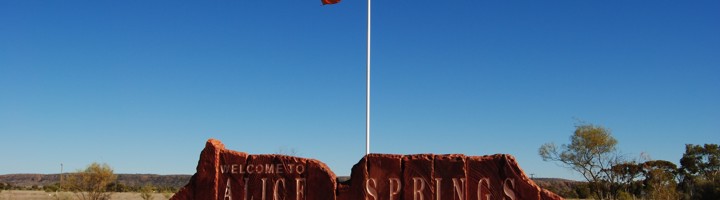Welkomstbord van Alice Springs in de rode outback van Australië, met vlaggenmast onder een blauwe lucht.