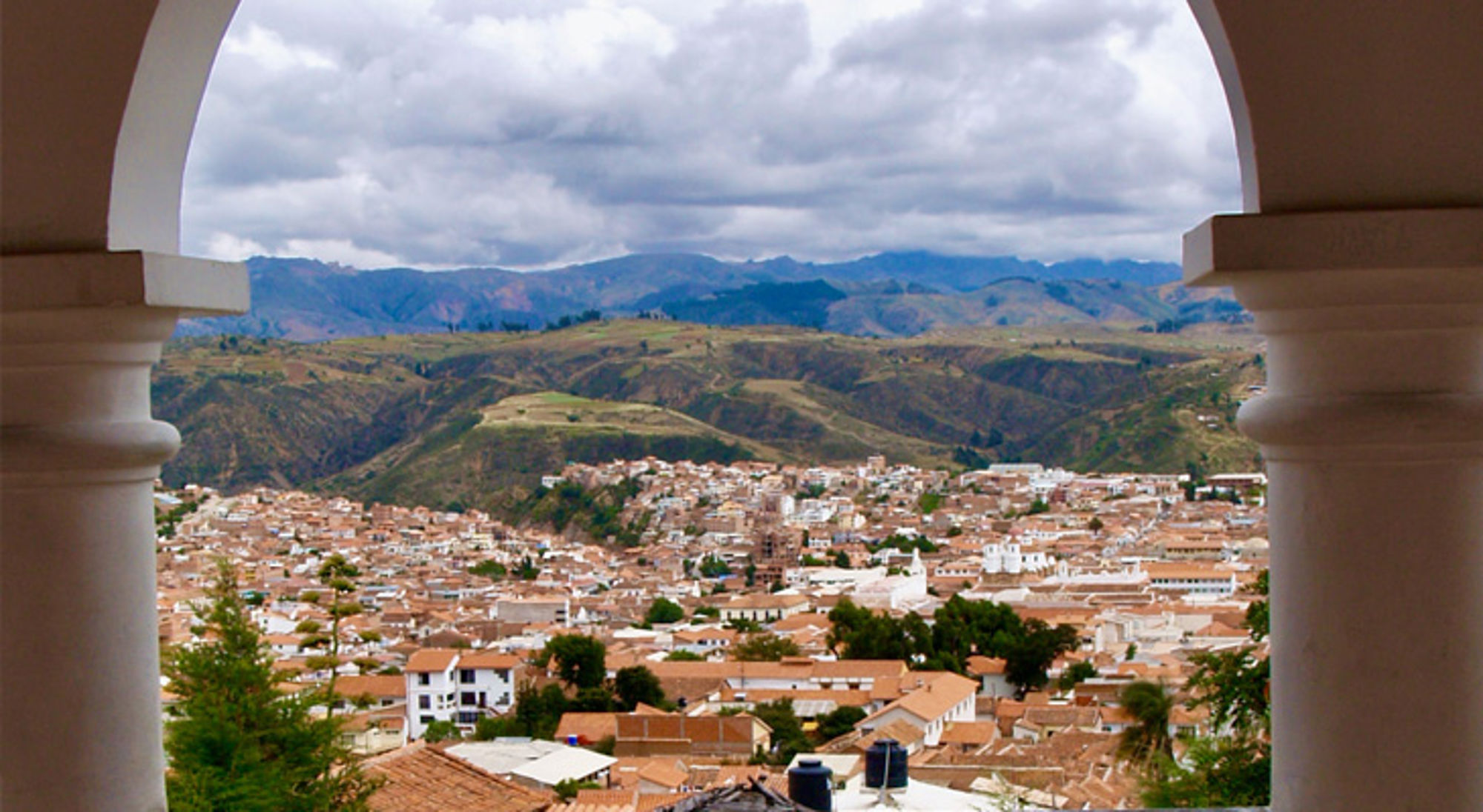 Uitzicht op de stad Sucre in Bolivia, met terracotta daken en groene bergen onder een bewolkte lucht.
