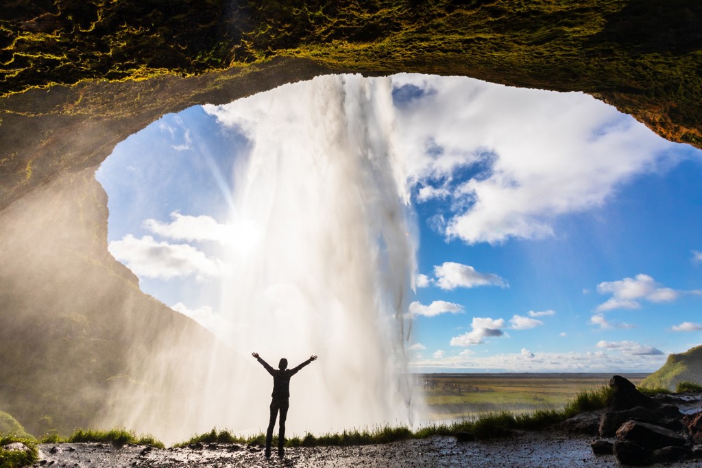 Reiziger met opgeheven armen achter een waterval in IJsland tijdens een 22-35ers groepsreis.