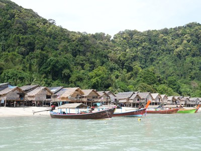 Longtailboten voor een strand met houten paalwoningen en groen regenwoud in Thailand.
