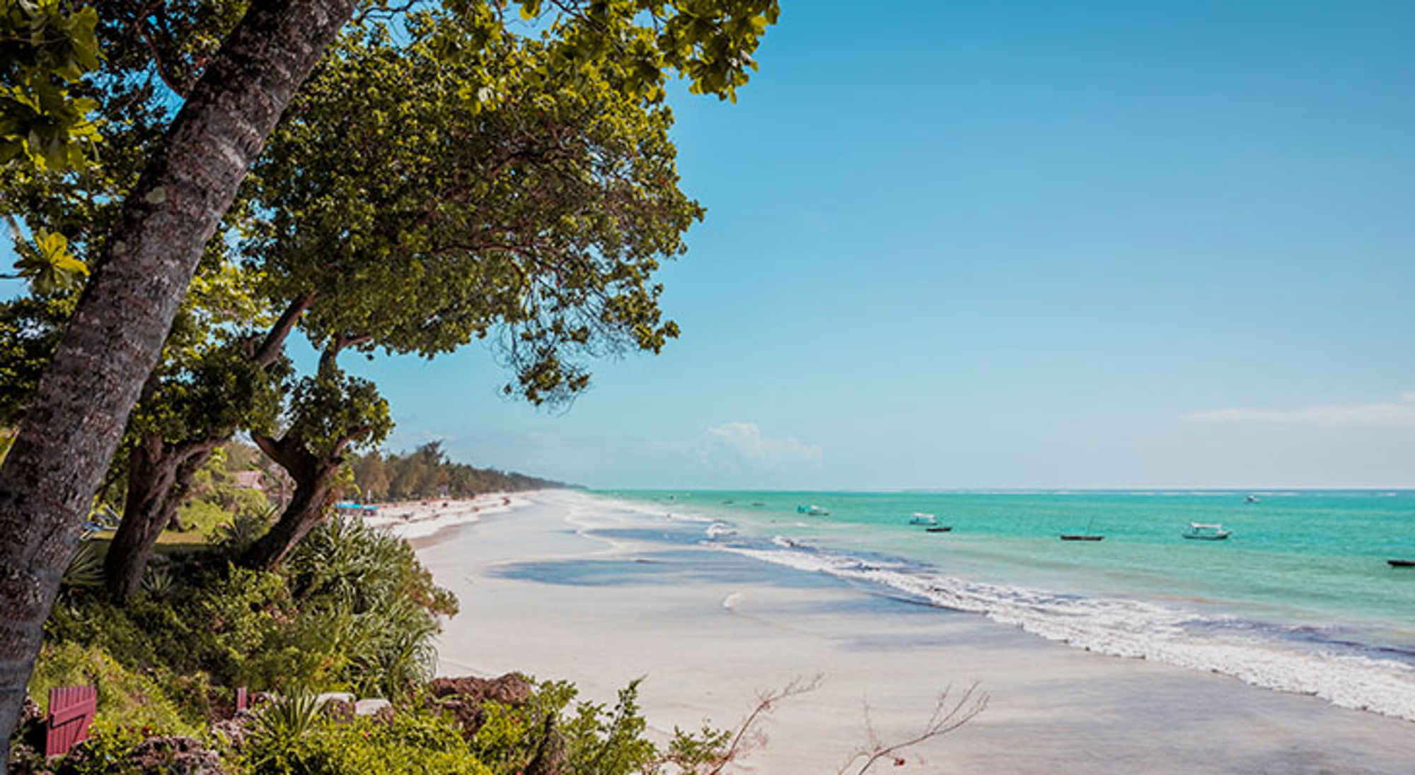 Wit zandstrand aan de Keniaanse kust met turquoise zee, palmbomen en bootjes voor de kust.