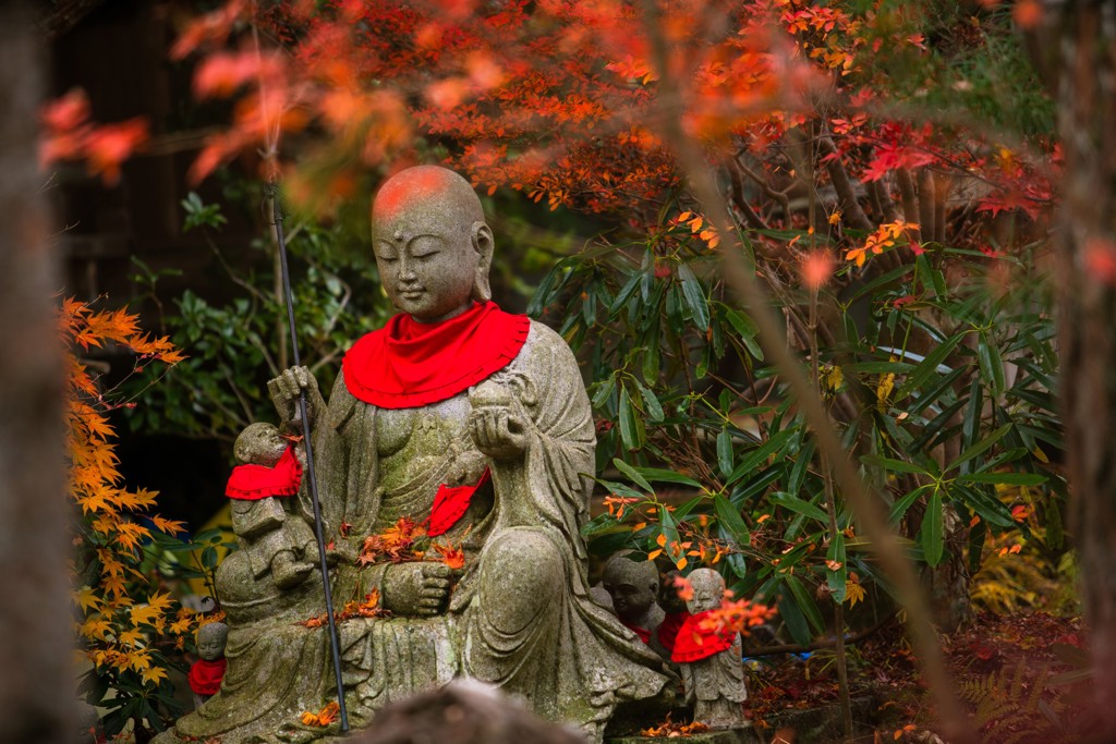 Jizo-beeld met rood slabbetje tussen herfstbladeren in een Japanse tempeltuin.