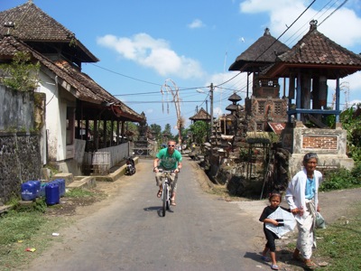 Fietser in een Balinees dorp met traditionele huizen en tempelgebouwen langs de straat tijdens de route Java, Bali, Lombok & Gili Air.