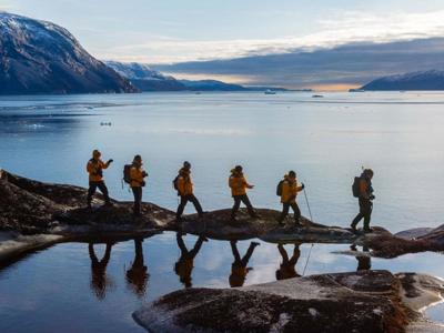 Wandelaars langs een fjordkust in Groenland, met bergen, ijs en spiegeling in het water.