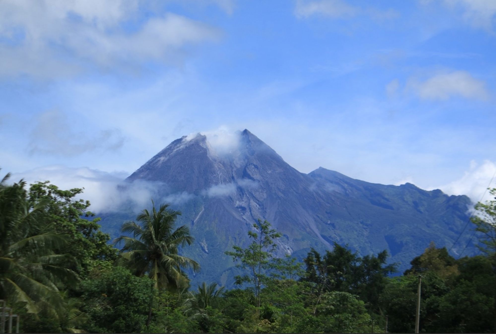 Vulkaan Mount Merapi in Indonesië met wolken rond de top en tropisch groen op de voorgrond.