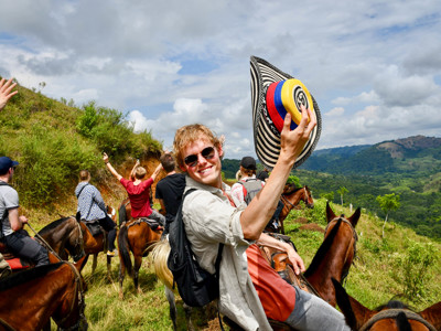Ruiters te paard in de groene bergen van Colombia, met reiziger die een kleurrijke sombrero vueltiao omhooghoudt.