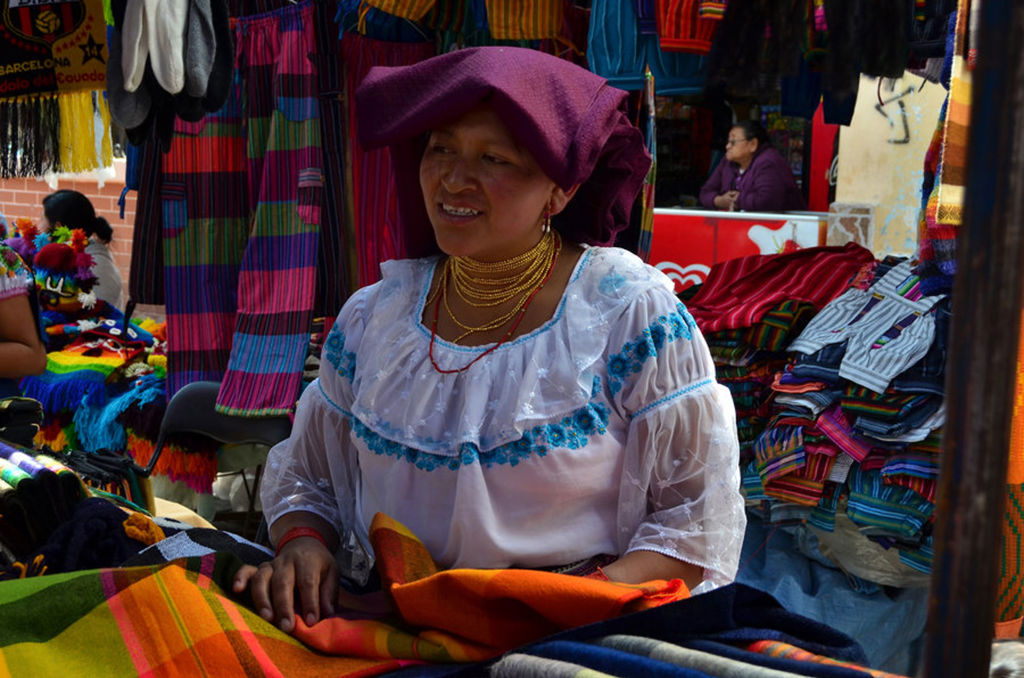 Marktvrouw in traditionele kleding tussen kleurrijke textielkramen in Ecuador tijdens een groepsrondreis.