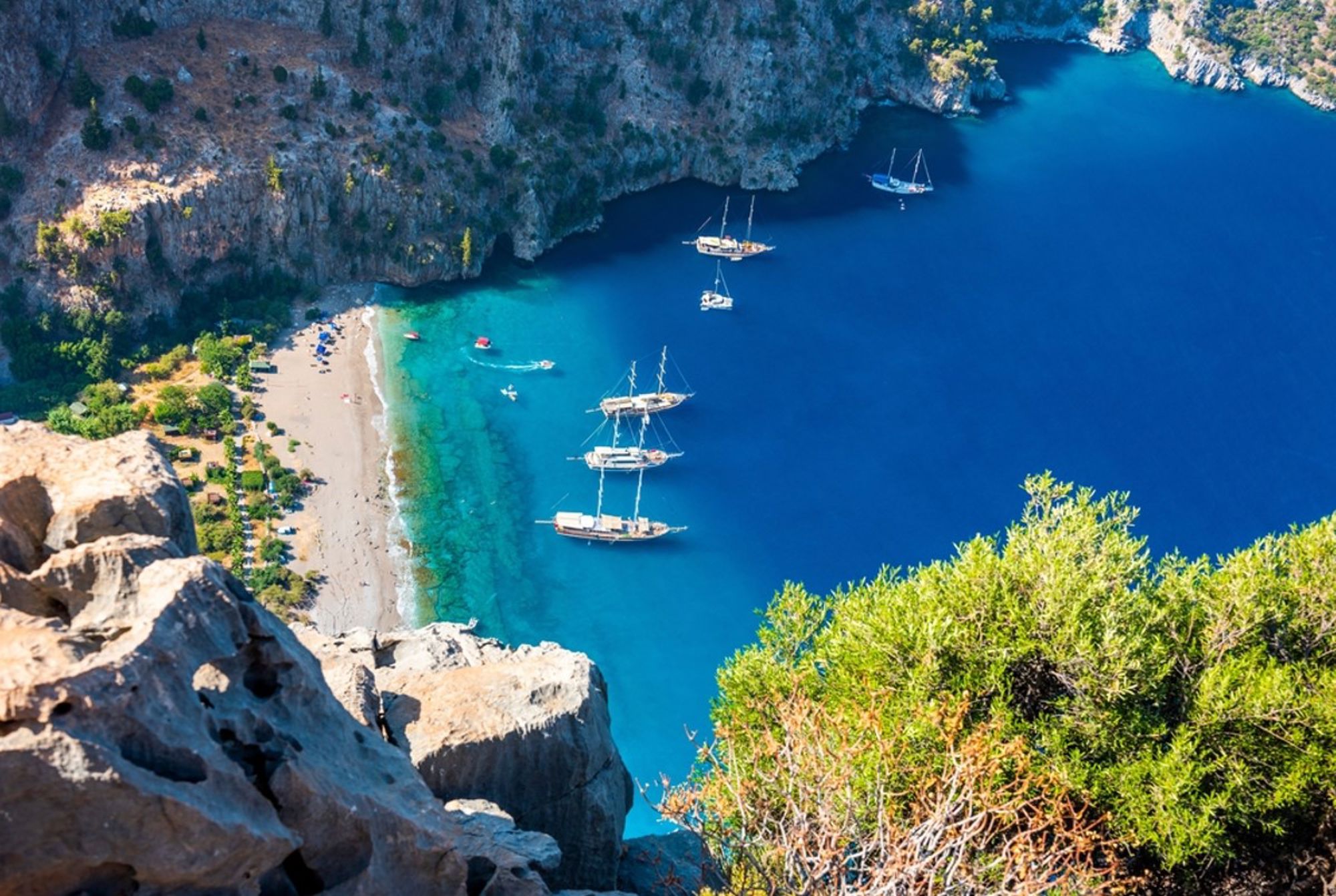 Uitzicht op een turquoise baai met strand en zeilboten tussen rotskliffen in Turkije.