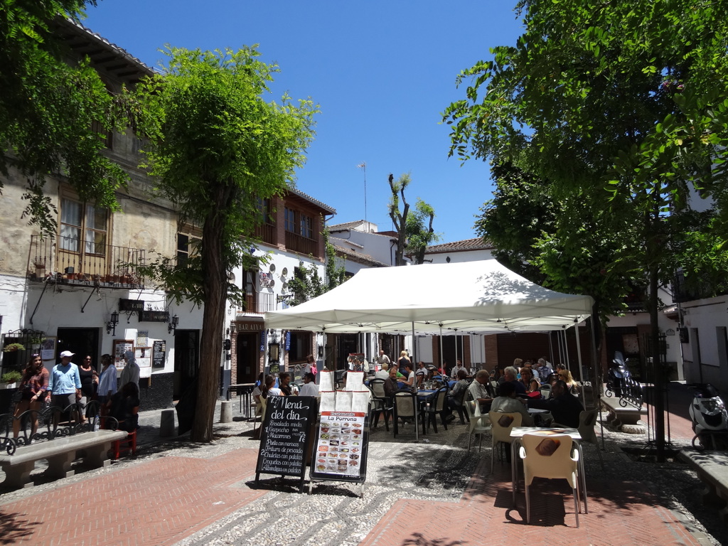 Terras op een sfeervol plein in Andalusië met witte huizen, bomen en gasten onder een grote luifel.
