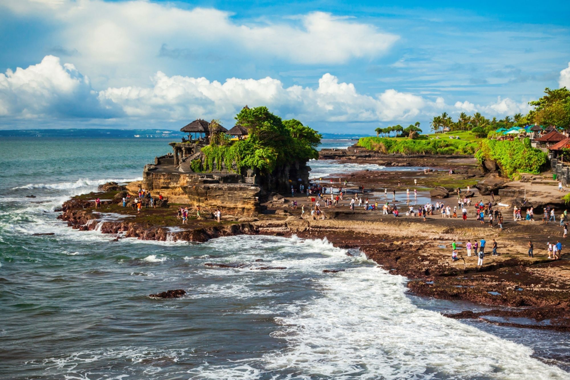 Tanah Lot-tempel op een rots aan de kust van Bali, met bezoekers op het strand en golven in de branding.