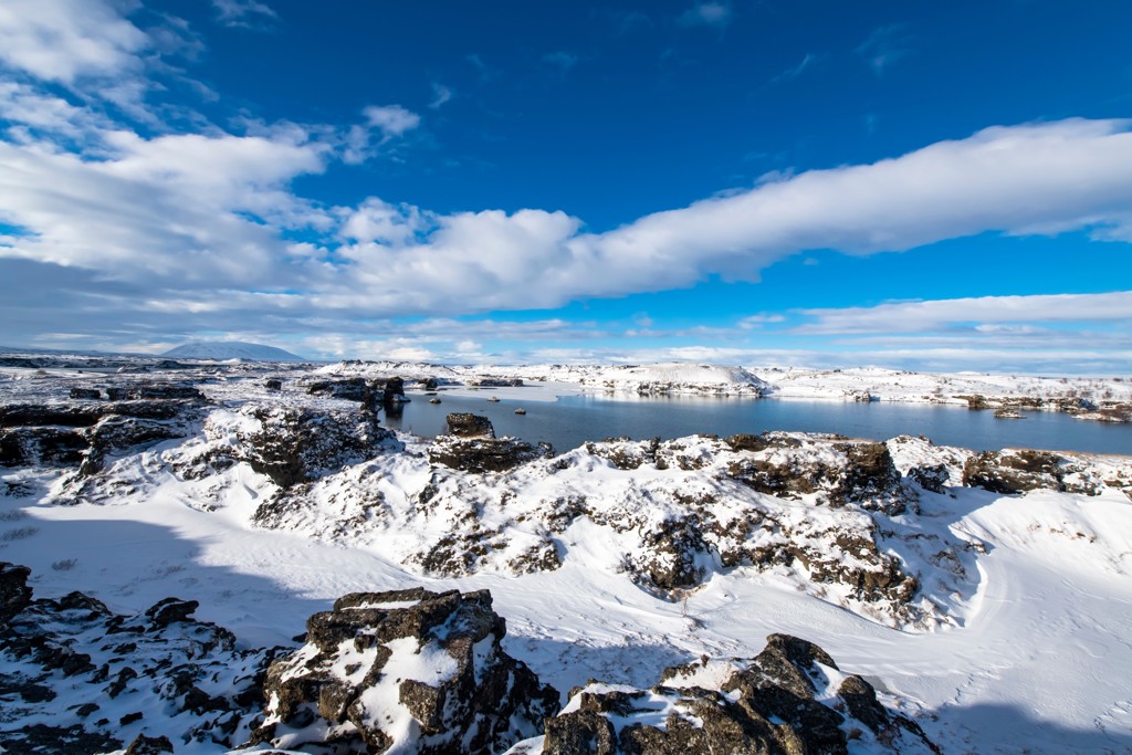 Sneeuwlandschap met lavavelden en helderblauw meer in Noord-IJsland, onder een uitgestrekte wolkenlucht.
