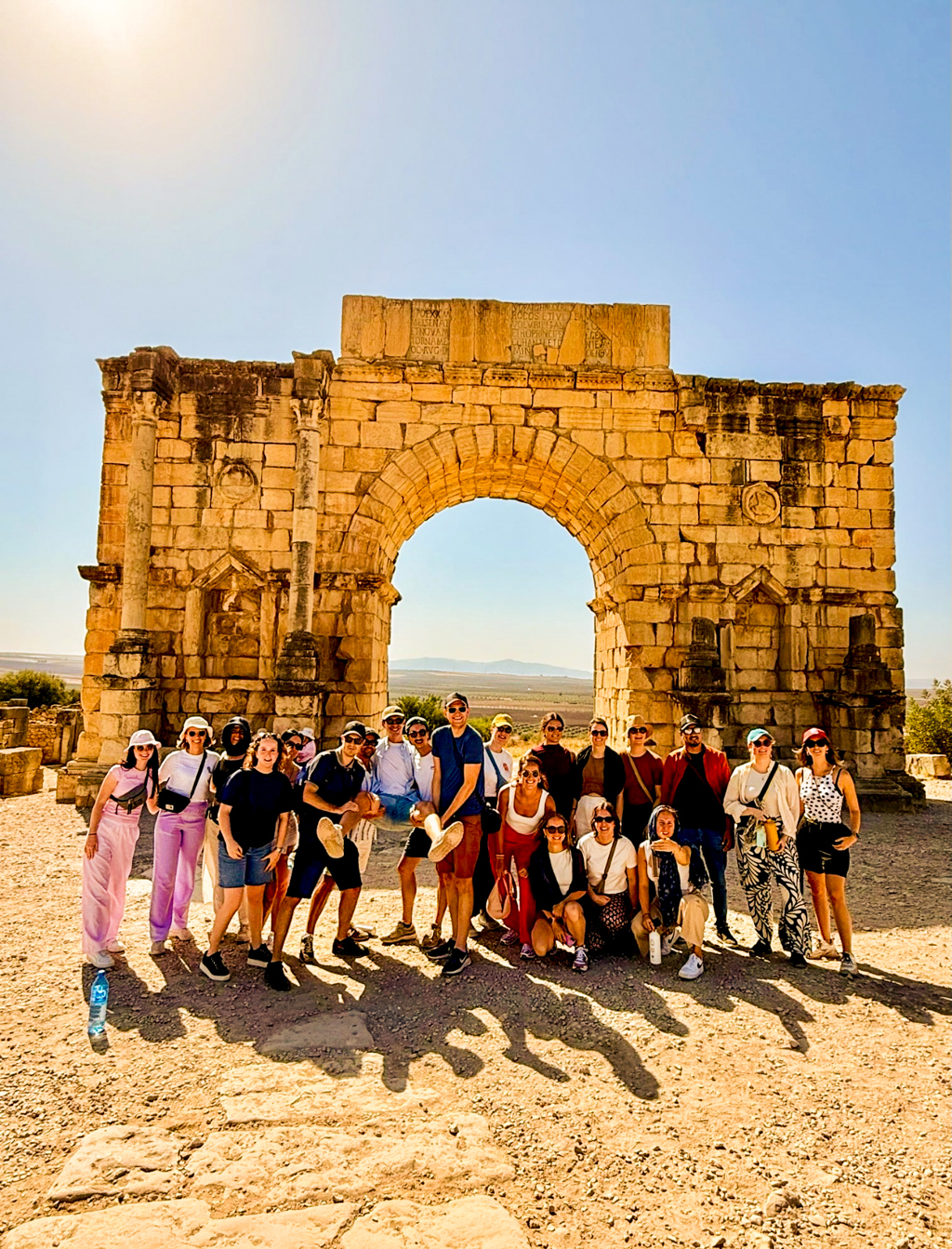 Groep 22-35ers reizigers poseert bij een Romeinse triomfboog tijdens rondreis door Marokko.