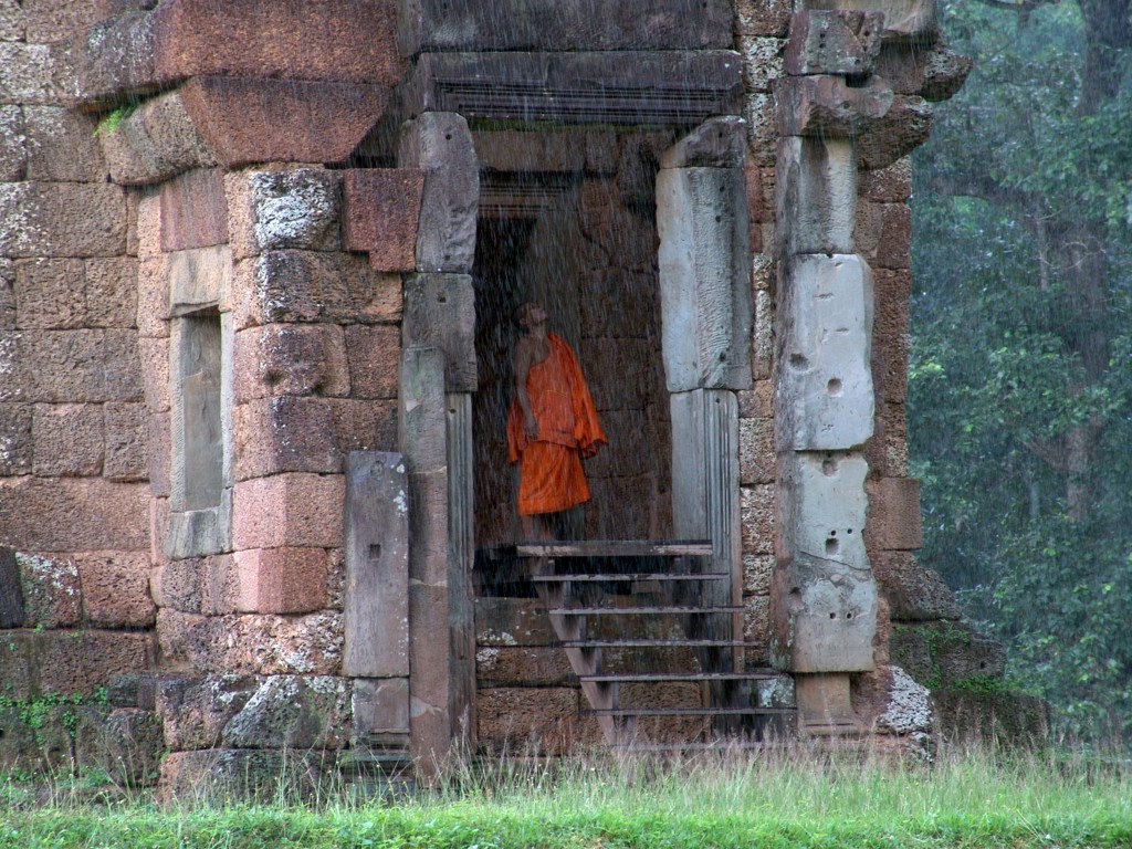 Boeddhistische monnik in oranje gewaad in de deuropening van een tempelruïne in Cambodja, tijdens regen.