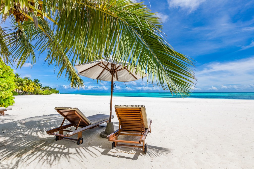 Strand op de Malediven met ligbedden onder een parasol en palmbomen aan een turquoise zee.