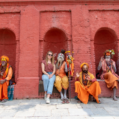 Reiziger tijdens 22-35ers reis Nepal poseert met kleurrijk geklede sadhu’s voor een rood tempelmuur.