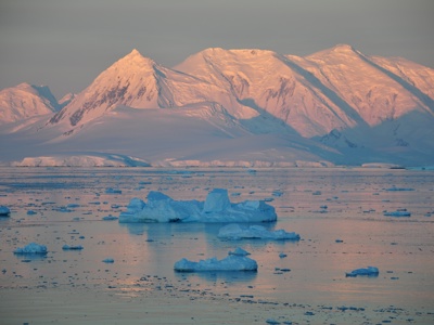 Besneeuwde bergketen langs de Antarctische kust met drijvende ijsbergen in zee bij zacht avondlicht.