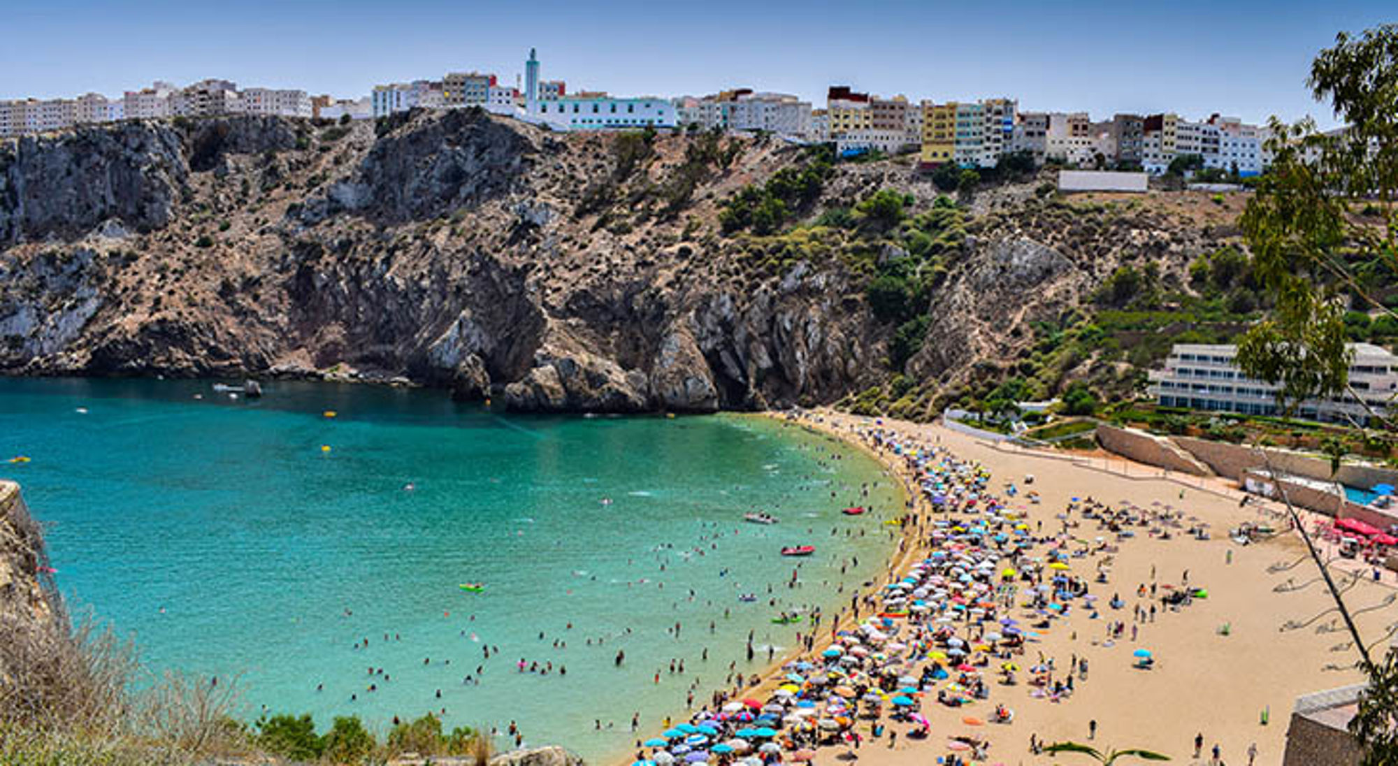 Druk strand in Al Hoceima, Marokko, met turquoise baai, rotsachtige kliffen en stad op de heuvel.