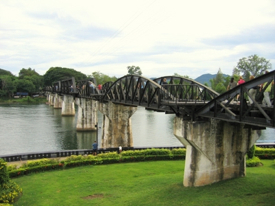 Brug over de rivier Kwai bij Kanchanaburi, Thailand, met wandelaars en groene oevers.