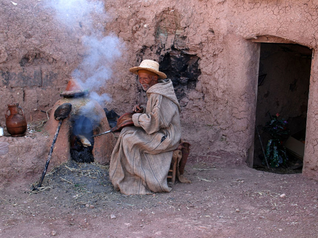 Man in traditionele djellaba bij lemen huis, zittend naast een rokende kleioven in Marokko.
