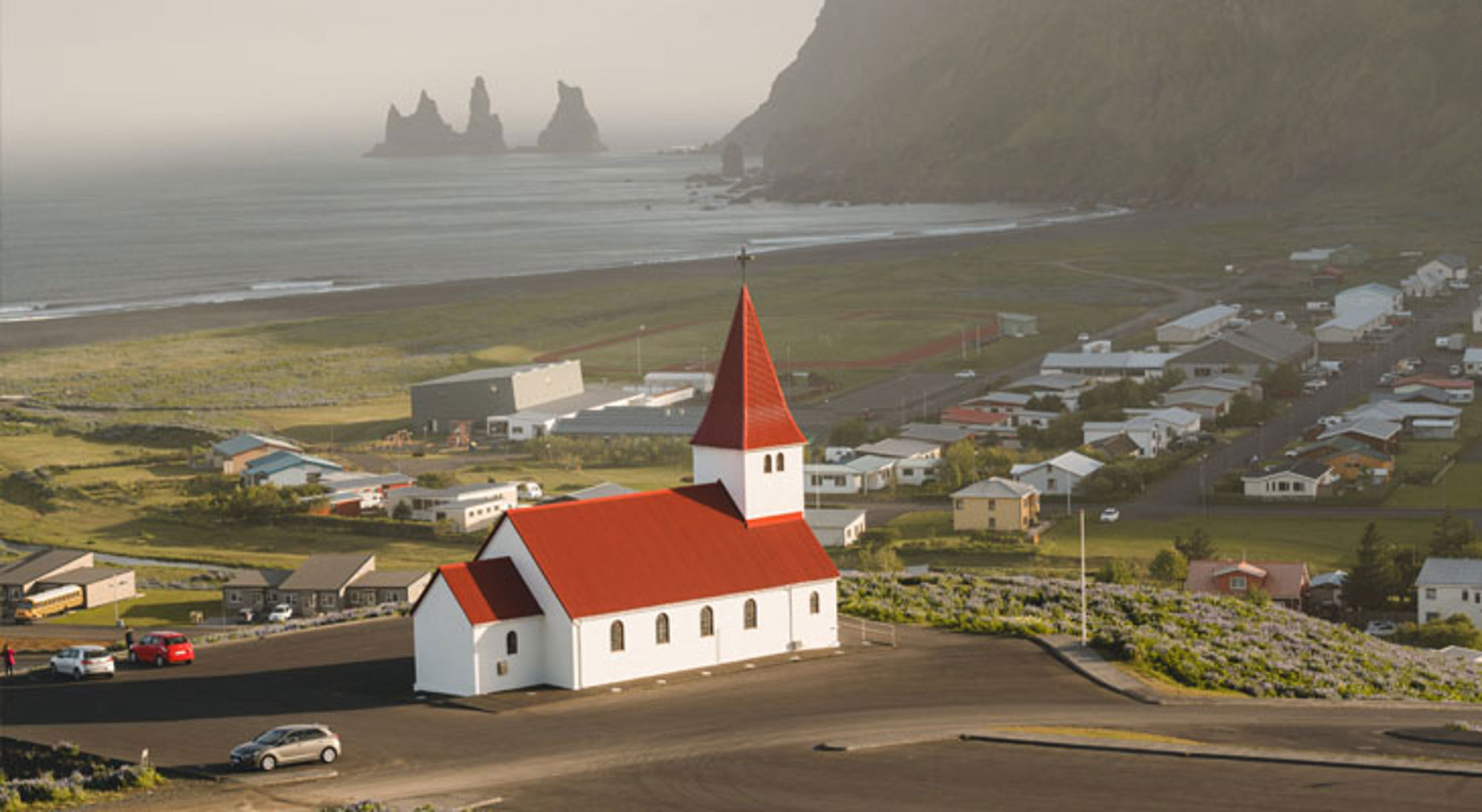 Witte kerk met rood dak in Vík í Mýrdal, met zwart zandstrand en Reynisdrangar rotsen op de achtergrond.