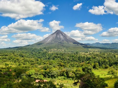 Vulkaan Arenal boven het groene regenwoud in Costa Rica, onder een blauwe lucht met wolken.