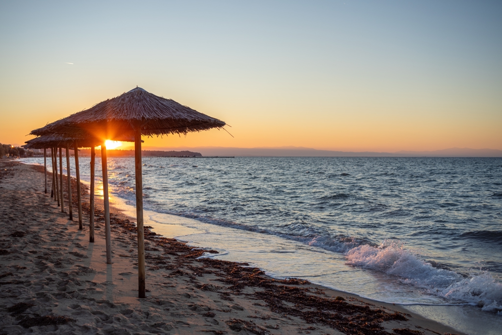 Zonsondergang op een strand in Noord-Griekenland met rieten parasols en rustige golven.