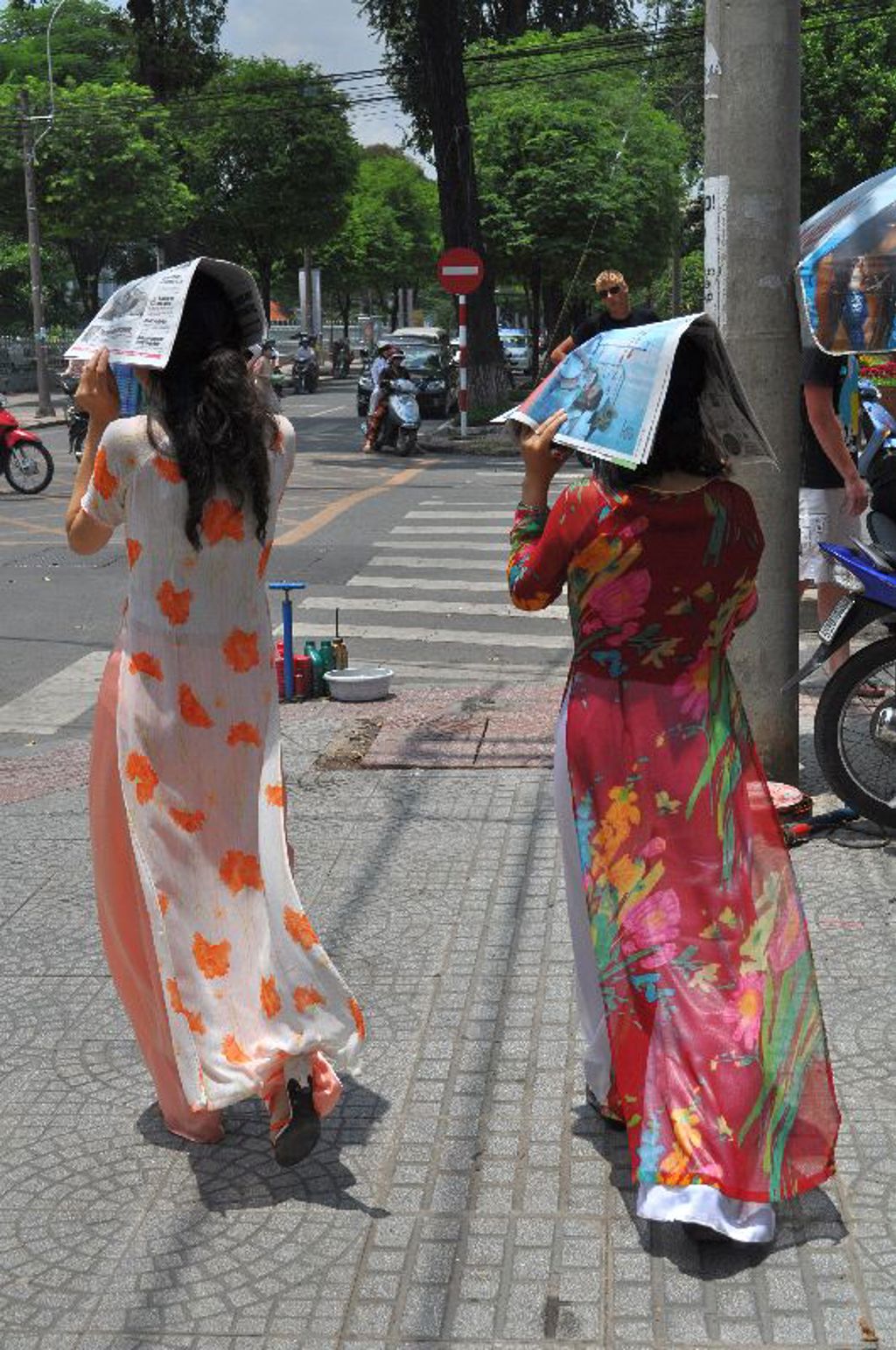 Twee vrouwen in áo dài steken een straat over in Vietnam en houden kranten boven hun hoofd tegen de zon.