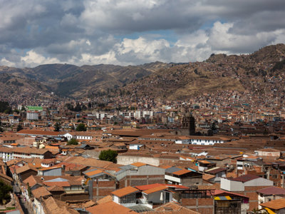 Panorama van Cusco met rode daken, kerk en omliggende Andesbergen onder een bewolkte lucht.