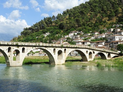 Oude stenen brug over de Osum-rivier bij Berat, met witte huizen tegen de groene heuvels in Albanië.
