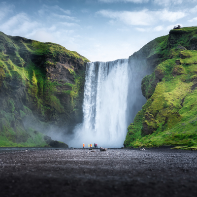 Skógafoss-waterval in IJsland met reizigers aan de voet, omringd door groene kliffen.