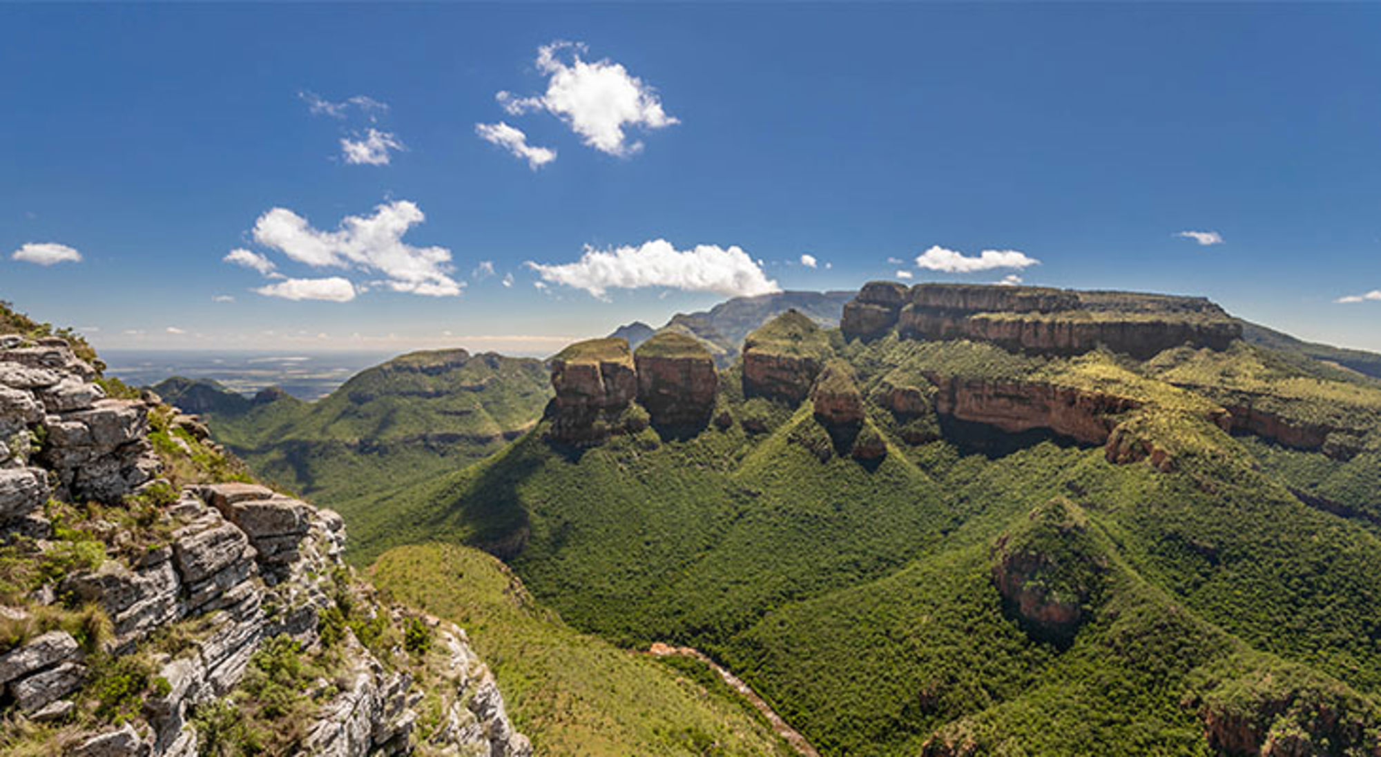Uitzicht op de Three Rondavels en Blyde River Canyon in Zuid-Afrika, met groene bergen en rotsformaties onder blauwe lucht.