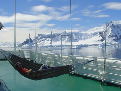 Reiziger ontspant in een hangmat op het dek tijdens een Spitsbergen-cruise, met ijsbergen en besneeuwde bergen op de achtergrond.