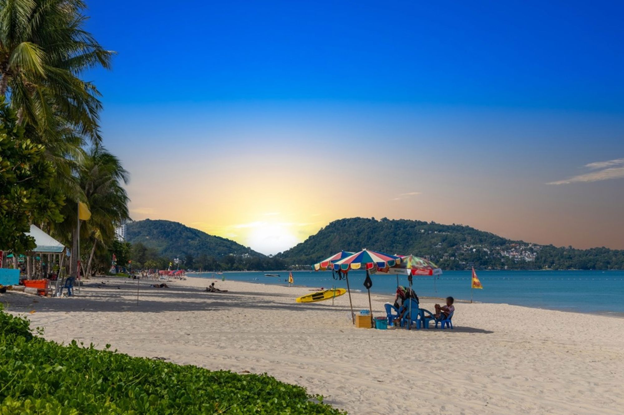 Zandstrand in Thailand bij zonsondergang, met palmbomen, parasols en uitzicht op zee en groene heuvels.