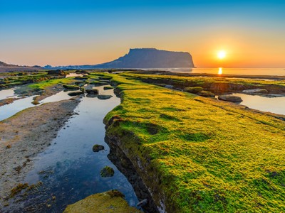 Zonsopgang bij Seongsan Ilchulbong op Jeju-do, met groene rotsen en getijdenpoelen langs de kust.