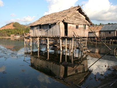 Houten paalwoning aan het water op de Kleine Sunda eilanden, met weerspiegeling en heuvels op de achtergrond.