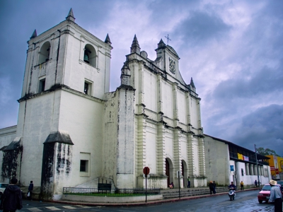 Witte kerk met klokkentoren in Guatemala, langs een straat onder dreigende wolken.