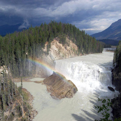 Waterval in de Canadese Rockies met regenboog boven een rivier, omringd door dennenbossen en bergen.