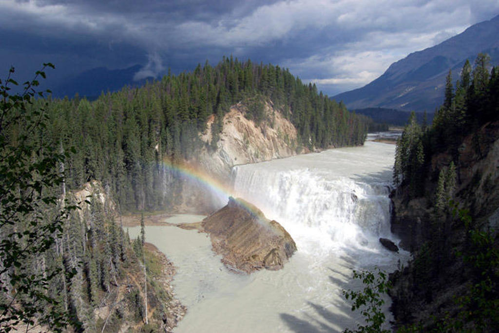 Waterval in de Canadese Rockies met regenboog boven een rivier, omringd door dennenbossen en bergen.