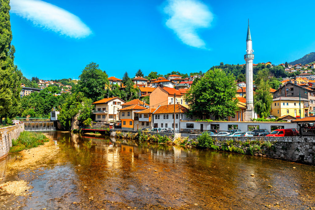 Stadsgezicht in de Balkan met rivier, huizen met rode daken en slanke minaret onder blauwe lucht.