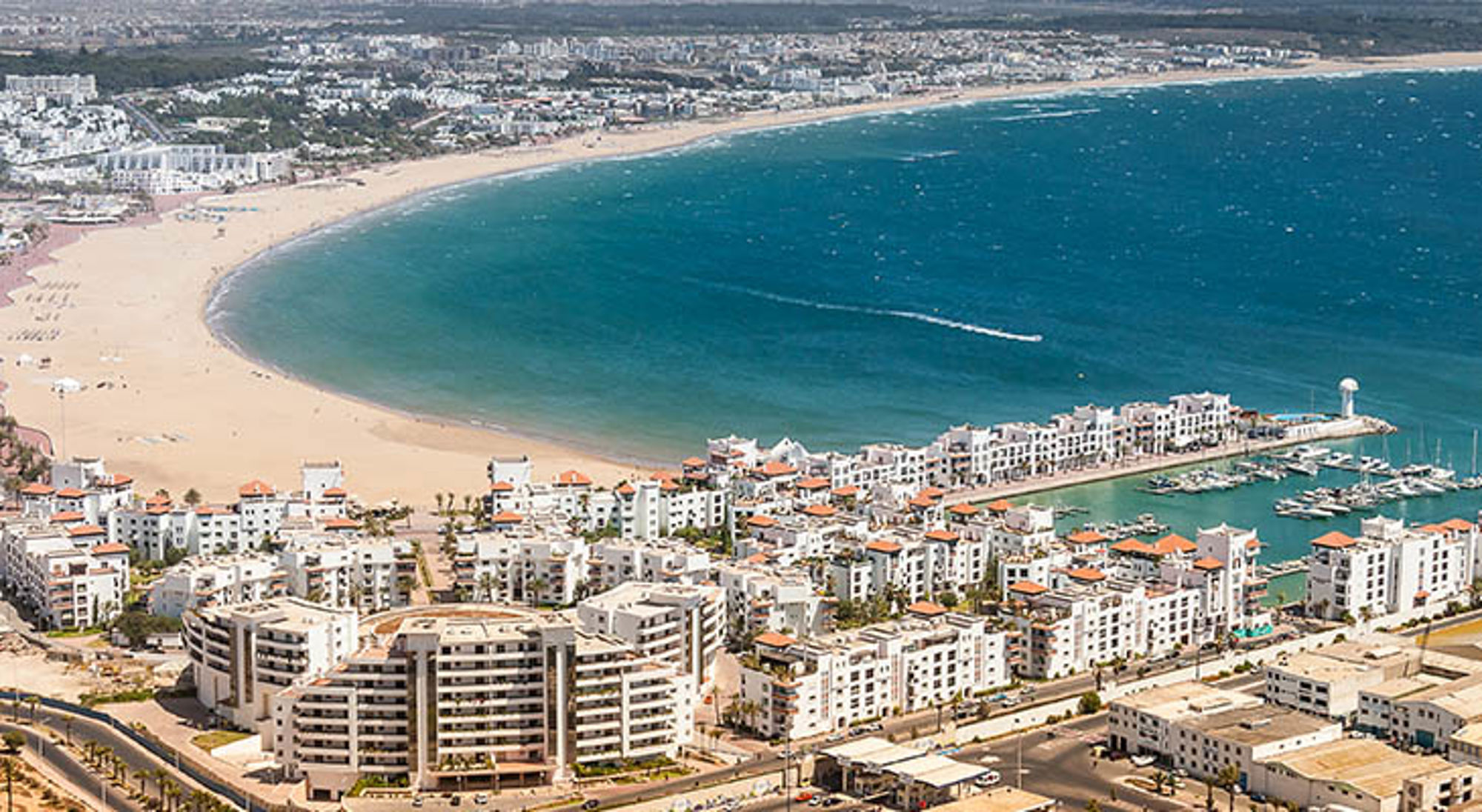 Agadir met brede zandbaai, blauwe Atlantische Oceaan en jachthaven met witte gebouwen.
