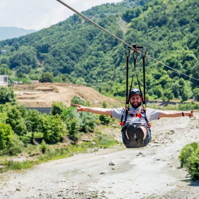 Man ziplinet bij Tirana over een rivierbedding met groene heuvels op de achtergrond.