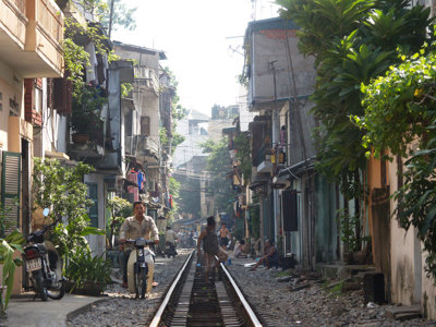 Smal straatje in Vietnam met spoorrails door de wijk, bewoners en scooters langs de huizen.