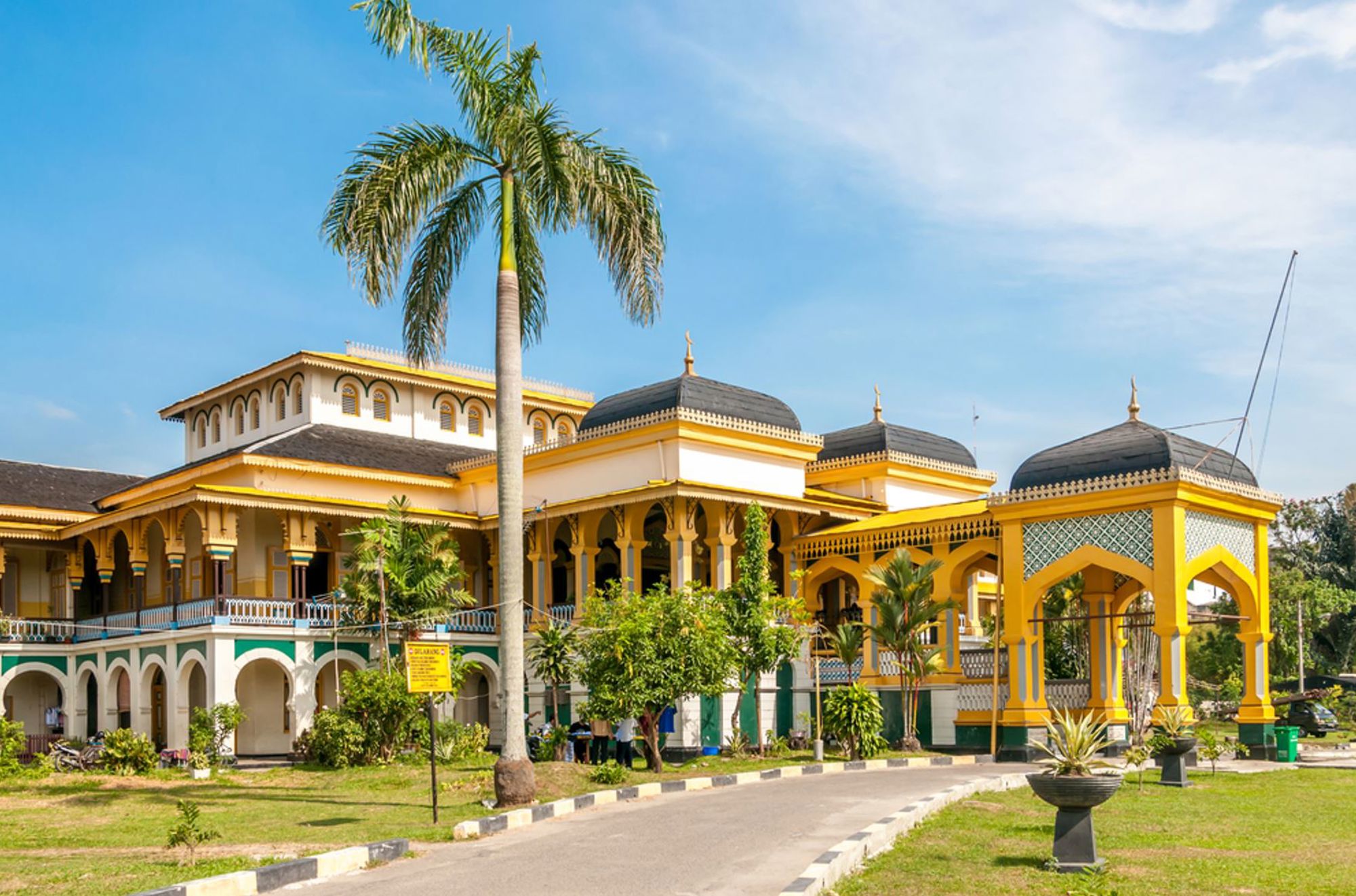 Geel paleis met zwarte koepels en palmbomen in Indonesië, onder een heldere blauwe lucht.