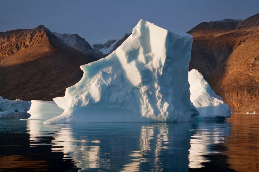 Grote ijsberg drijft in kalm water bij rotsachtige kust, tijdens groepsrondreis Spitsbergen, Groenland en IJsland.