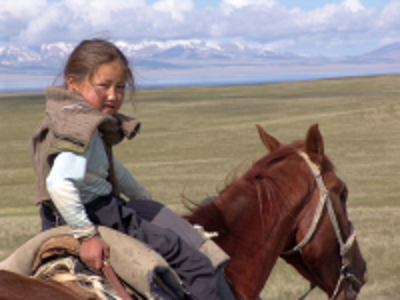 Kind op een paard in de uitgestrekte steppe van Kirgistan, met besneeuwde bergen op de achtergrond.