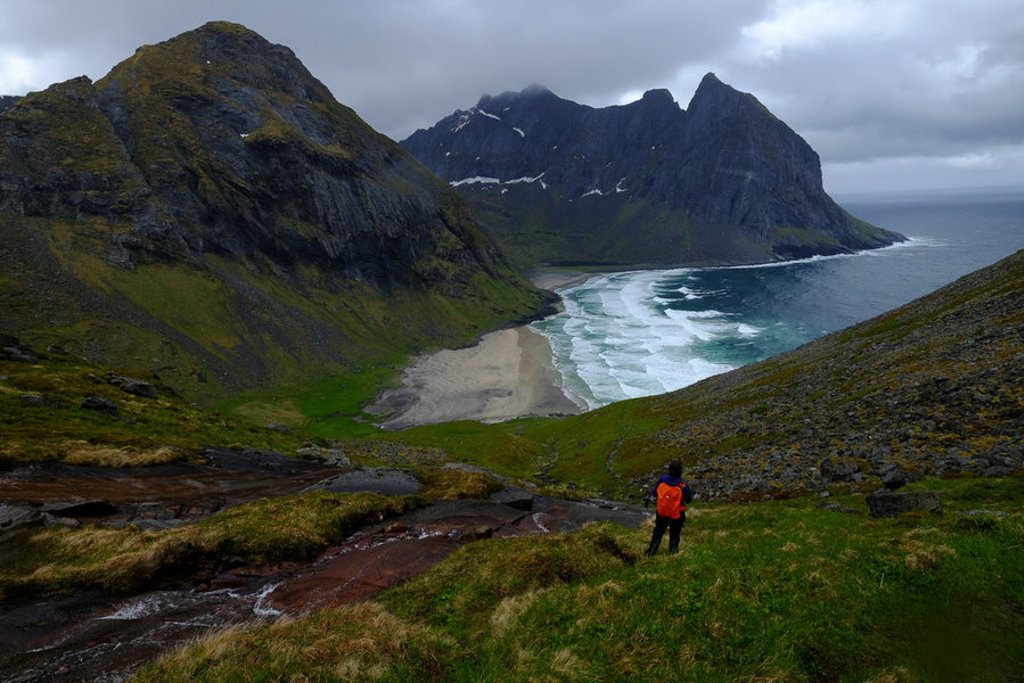 Hiker met rode rugzak kijkt uit over bergkust en zandstrand op de Lofoten, Noorwegen.